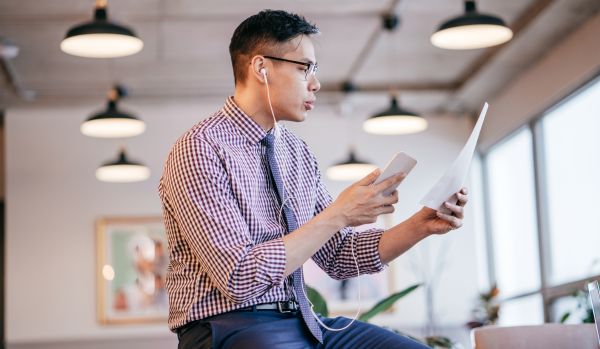 A man is sitting on a table. He's on the phone with headphones plugged in, reading a sheet of paper as he talks.