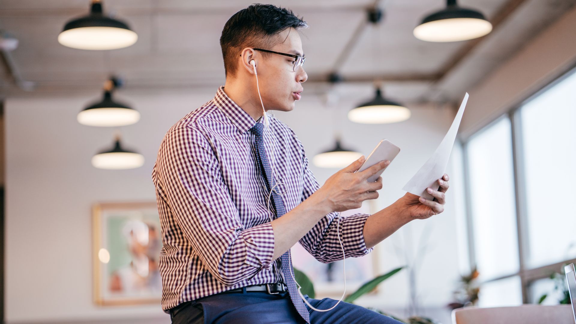 A man is sitting on a table. He's on the phone with headphones plugged in, reading a sheet of paper as he talks.
