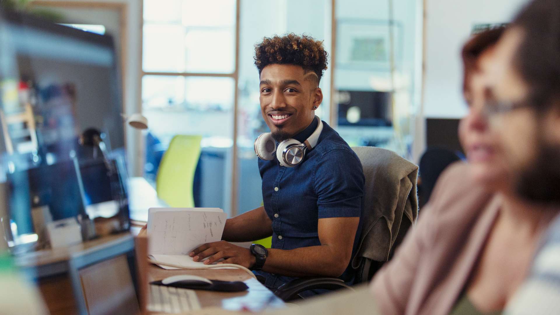 A Gen Z man smiles at the camera while opening his notebook in an office setting.