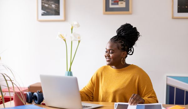 A young woman in a yellow shirt is working on her laptop in her home office.