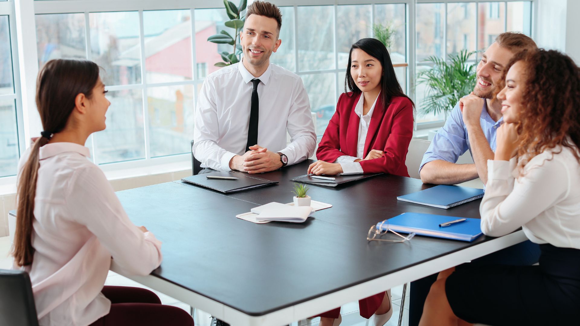 There is a woman sitting across from four people who are interviewing her. They are smiling and have notebooks with pens on the table in front of them.
