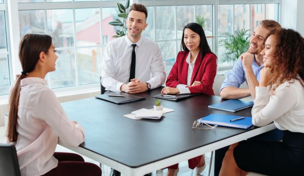 There is a woman sitting across from four people who are interviewing her. They are smiling and have notebooks with pens on the table in front of them.