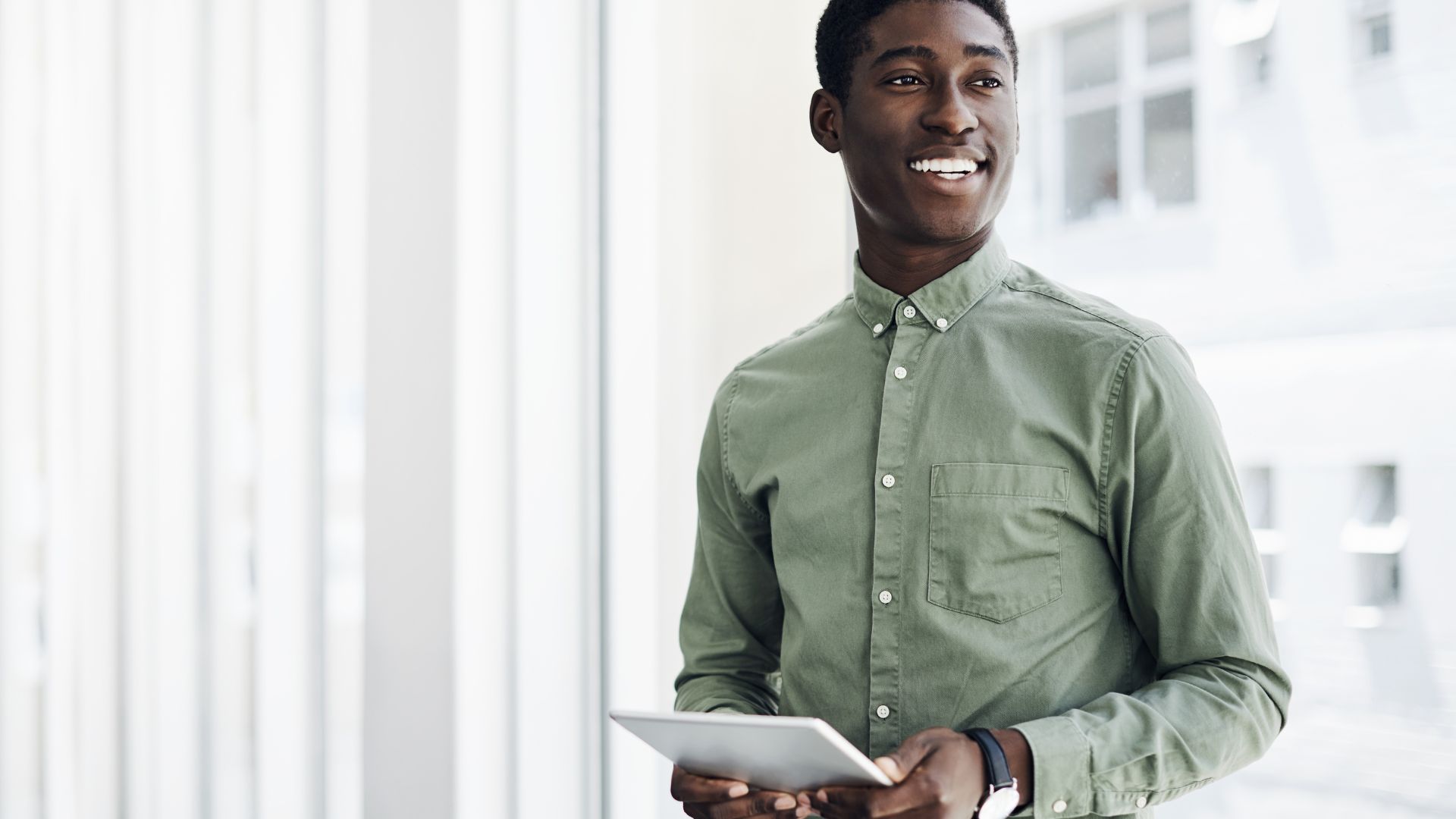 A young man is standing and holding a tablet, smiling off into the distance.