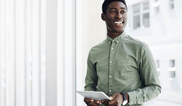A young man is standing and holding a tablet, smiling off into the distance.