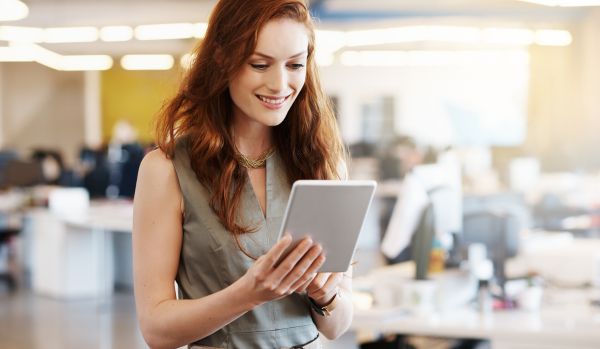 A woman is standing in an open office space, smiling at the tablet she's holding.