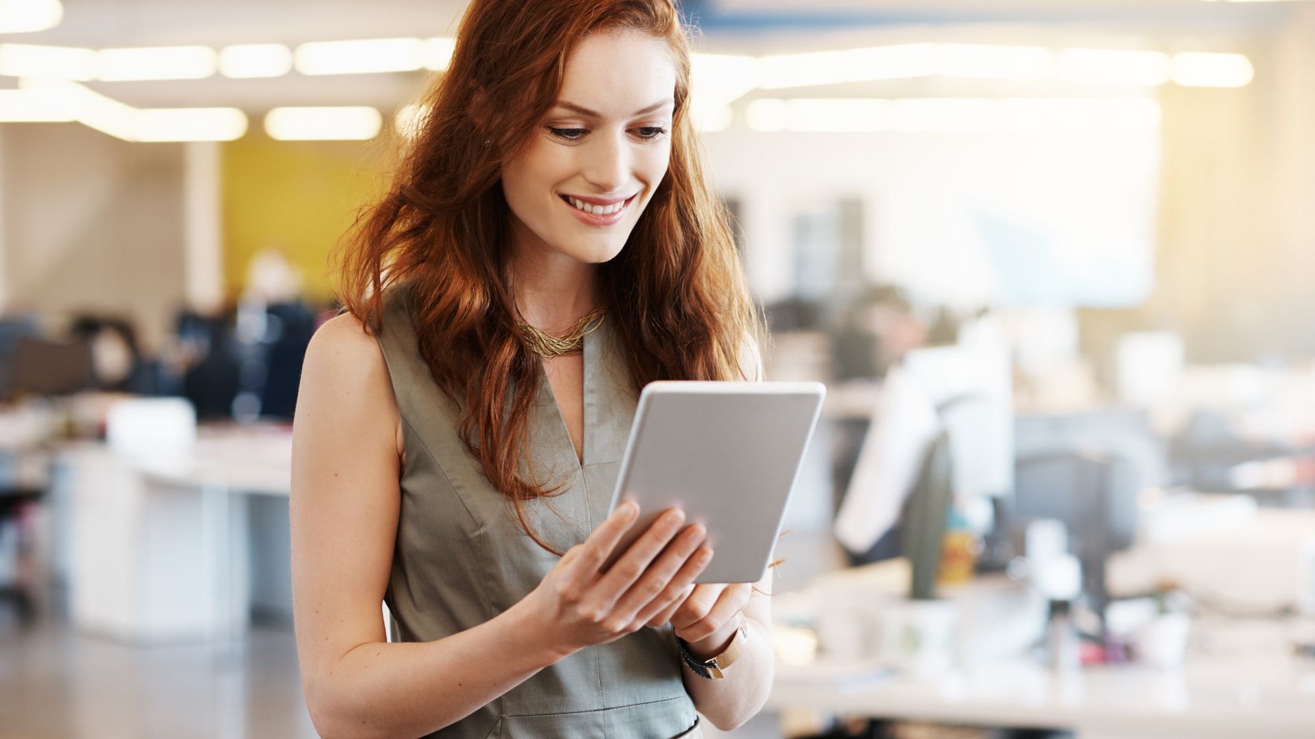 A woman is standing in an open office space, smiling at the tablet she's holding.