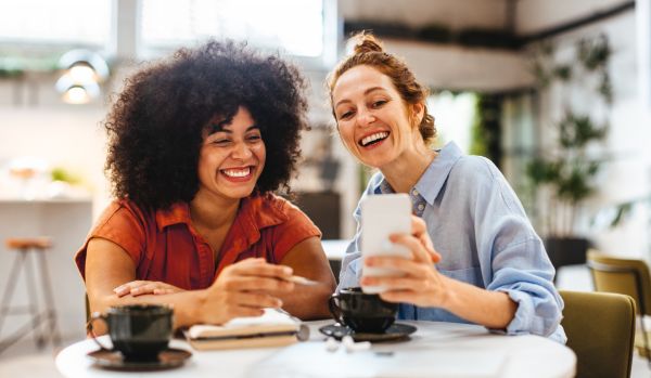 Two women are working in the break room/common area of their office. One is holding up her phone and they are both smiling at it.