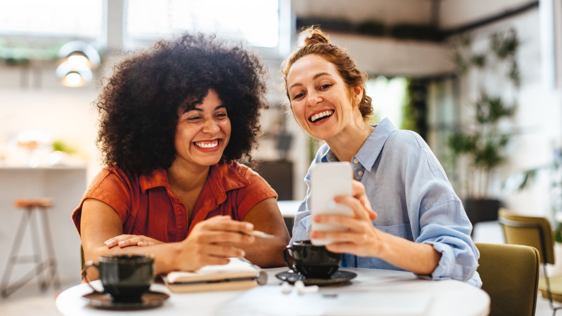 Two women are working in the break room/common area of their office. One is holding up her phone and they are both smiling at it.