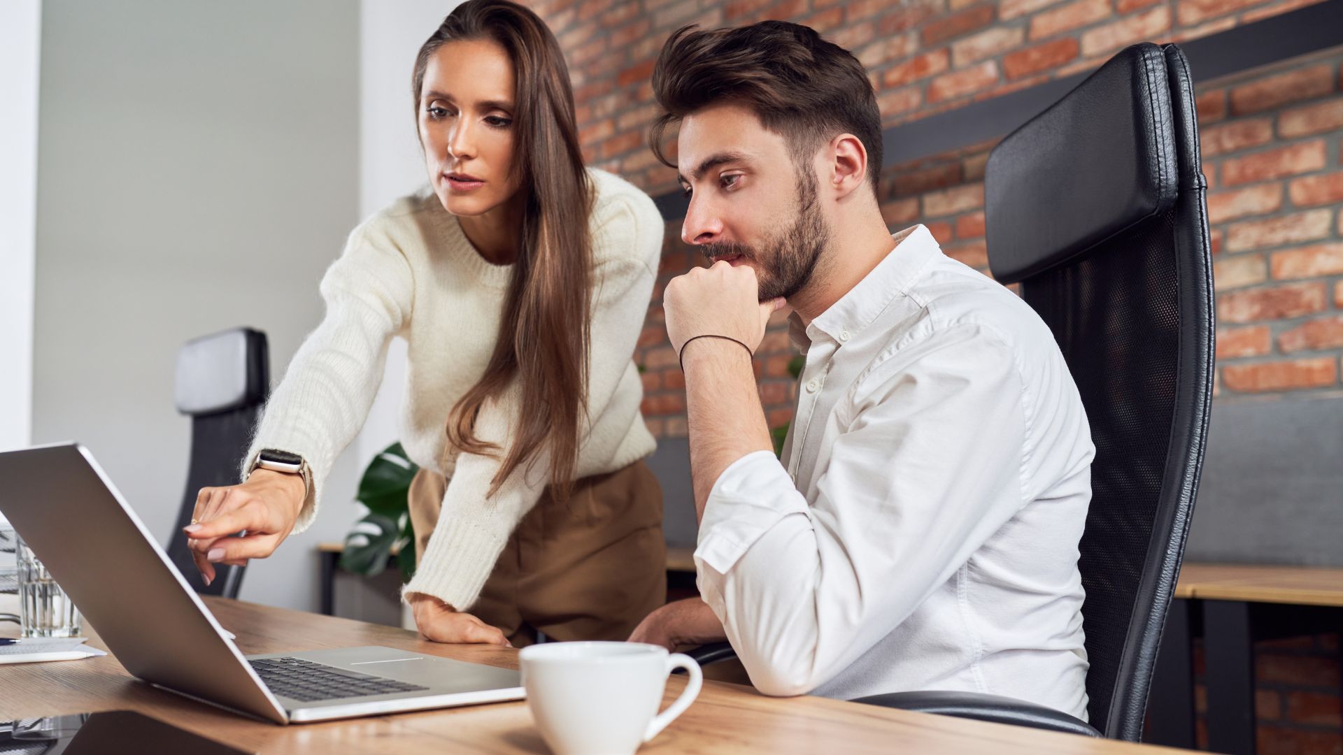 A woman is standing behind a colleague, pointing at his laptop and explaining something to him.