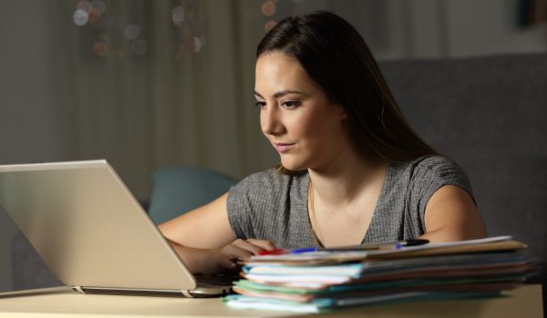 A woman is working on her laptop with a stack of paperwork next to her.