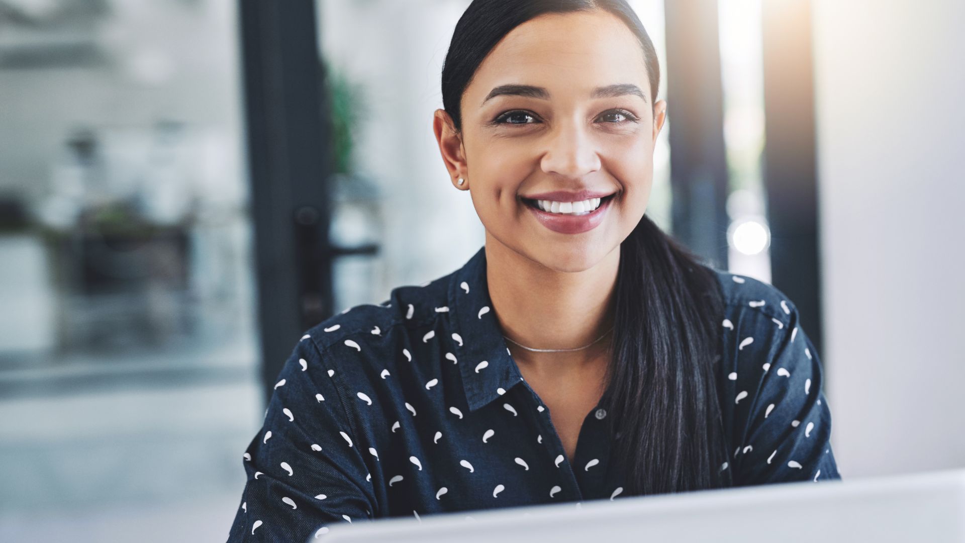 A young woman is smiling over the laptop at the camera.
