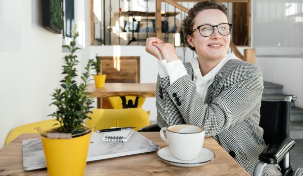 A woman in a wheelchair is sitting at a small table with her laptop and coffee, smiling into the distance.