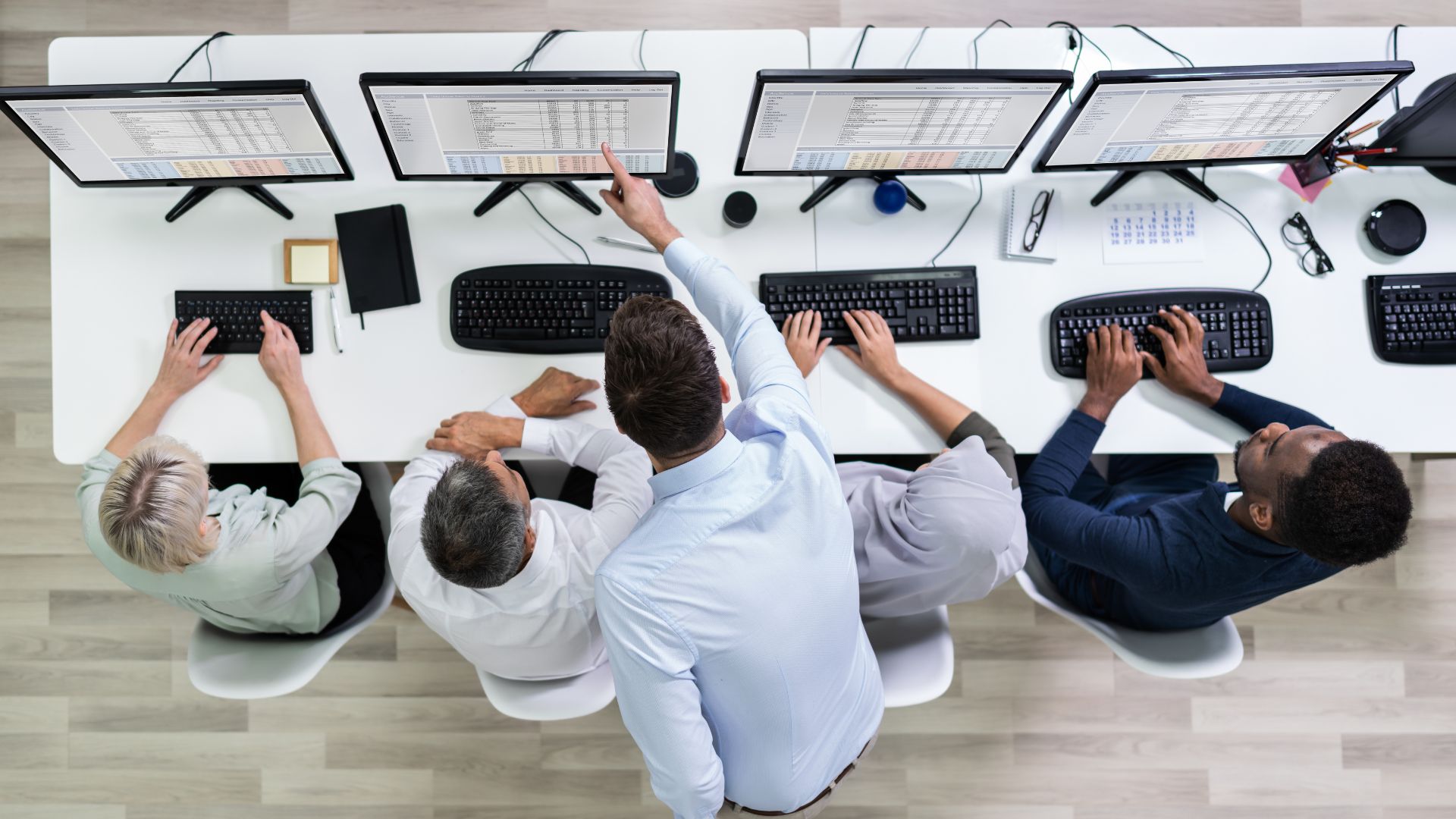 We are looking down from directly overhead at a man teaching 4 employees how to use an AI tool. The employees are seated in a row while the man is standing behind them, leaning over and pointing at the screen.