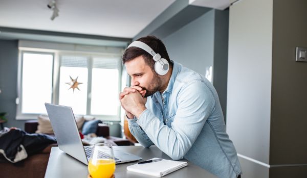 A young man is sitting at his kitchen counter participating in a live training on his laptop. He is wearing headphones and resting his head on his hands.