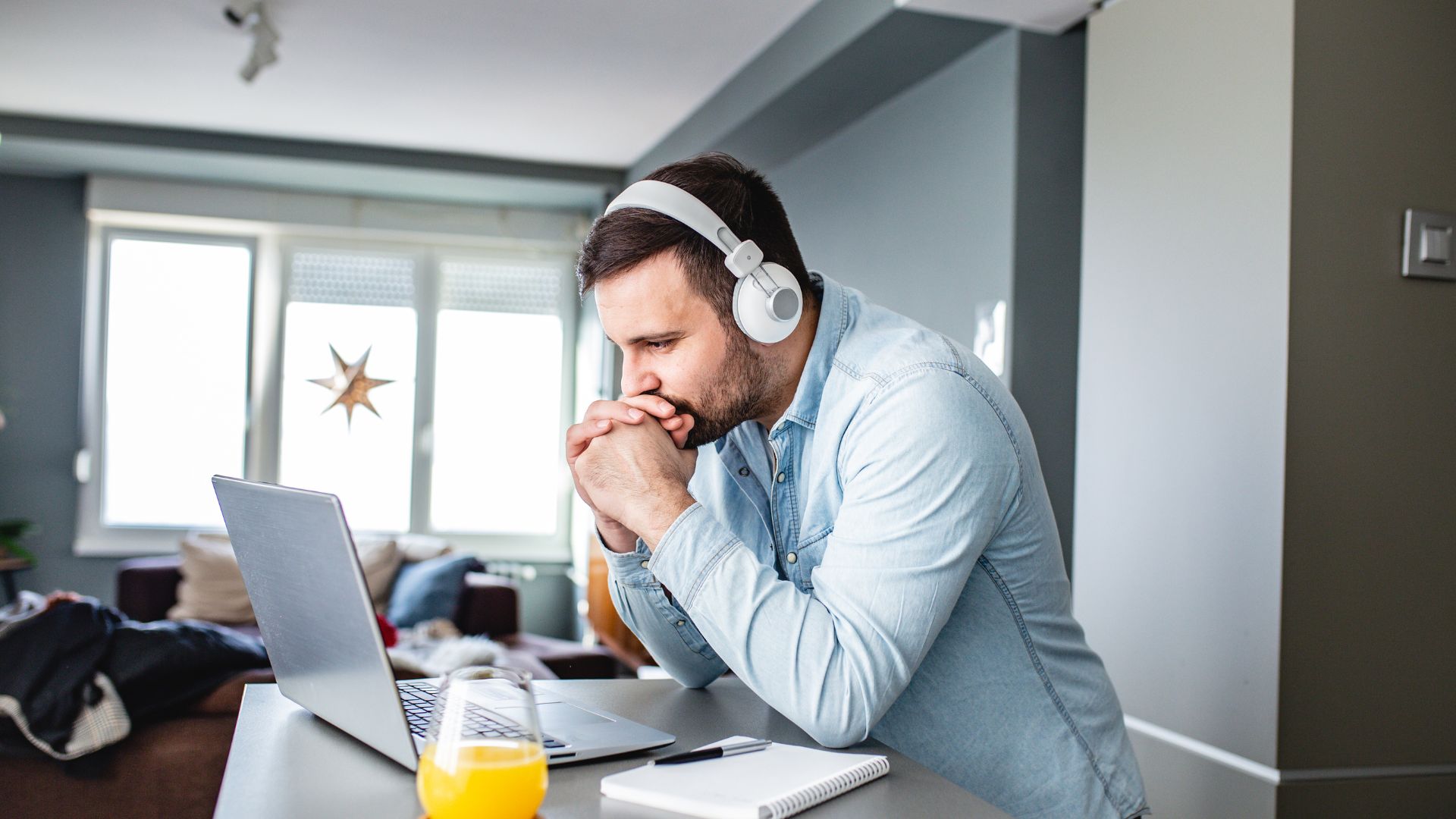 A young man is sitting at his kitchen counter participating in a live training on his laptop. He is wearing headphones and resting his head on his hands.