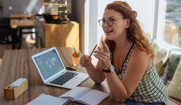 A young woman in glasses is working from a cafe on her laptop with a notebook.
