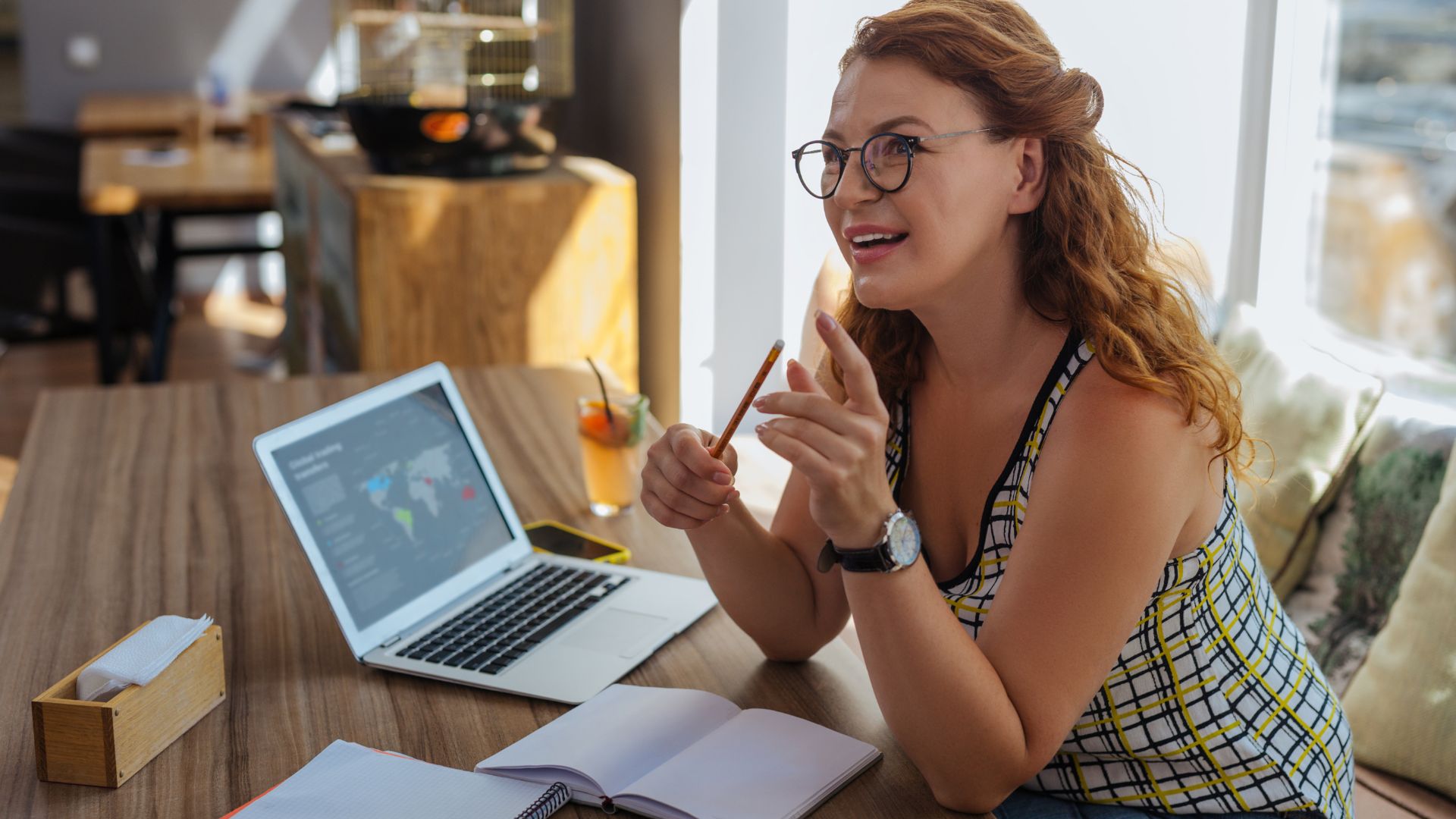 A young woman in glasses is working from a cafe on her laptop with a notebook.