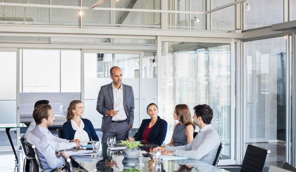 A man is standing at the head of a conference table, presenting to a diverse group of colleagues.