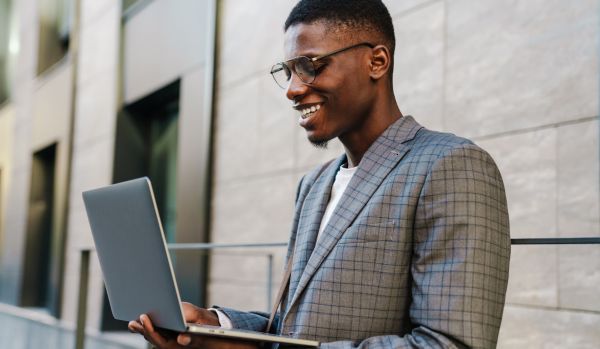 A young man wearing classes is standing outside holding his laptop open and smiling down at his screen.