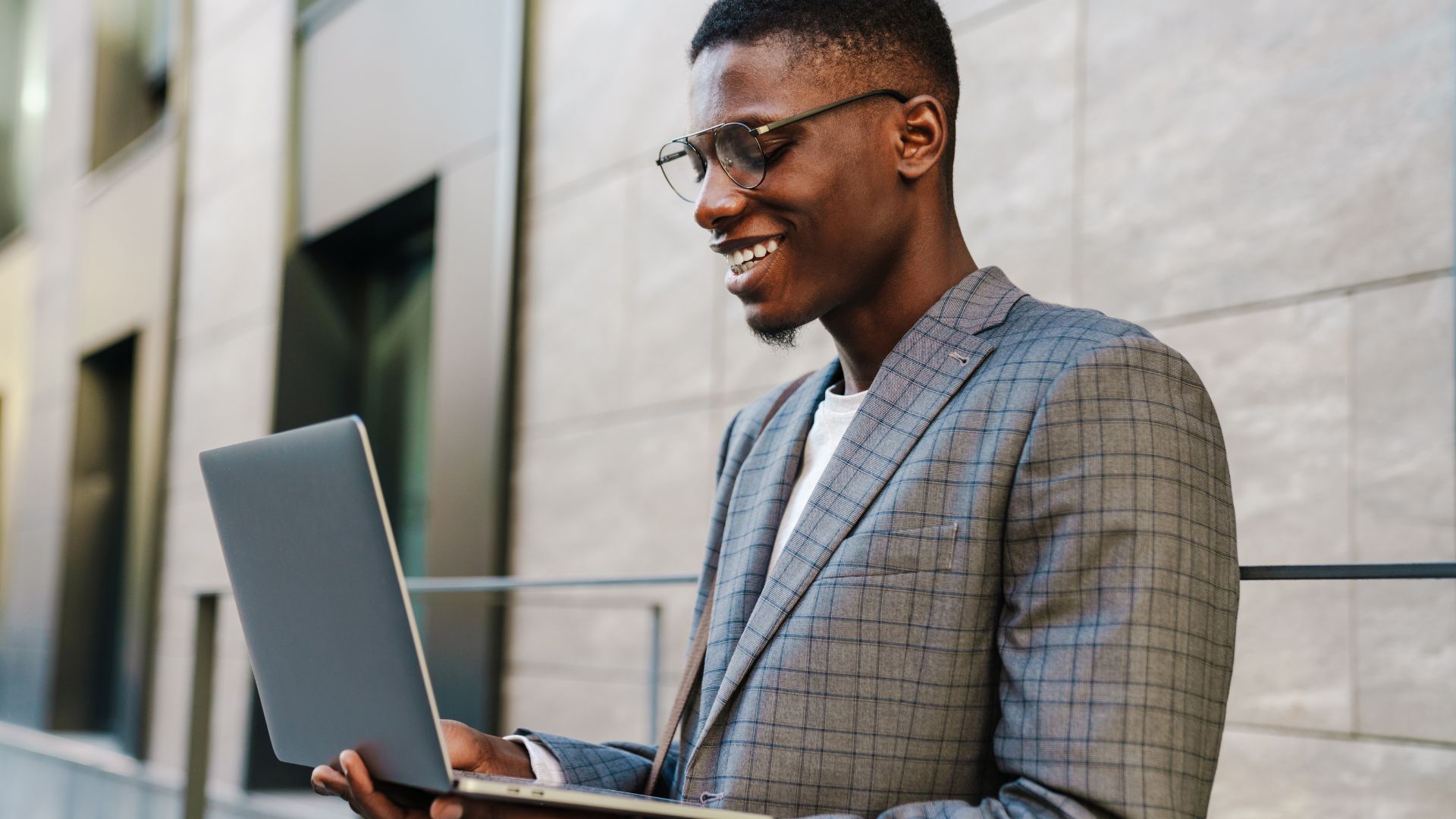 A young man wearing classes is standing outside holding his laptop open and smiling down at his screen.