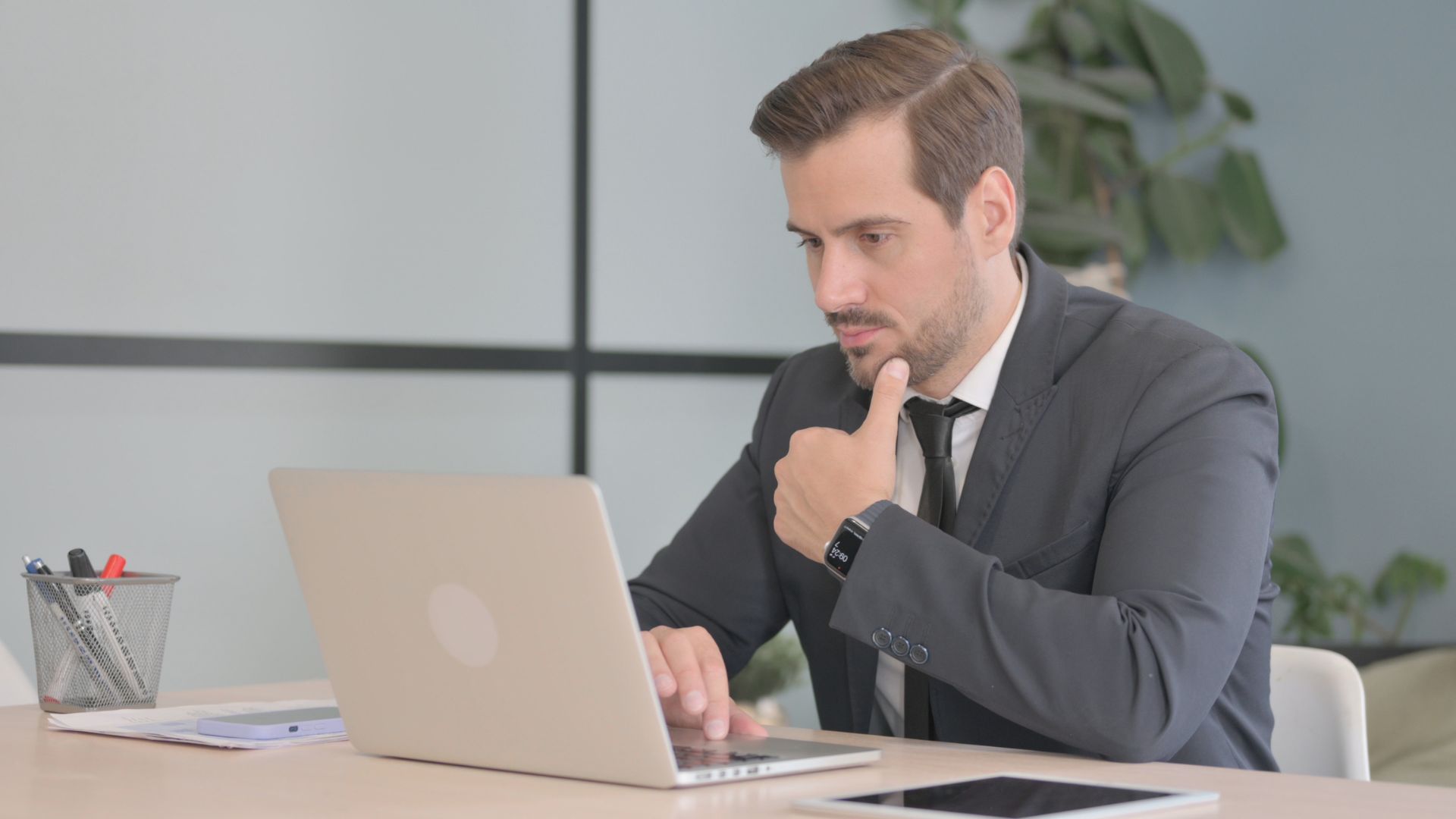A man in a suite is looking down at his laptop in his work office, thinking deeply about something.