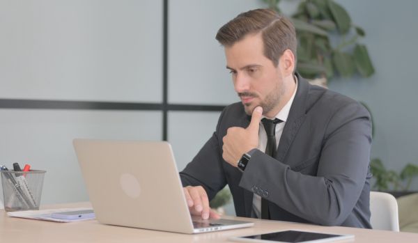 A man in a suite is looking down at his laptop in his work office, thinking deeply about something.
