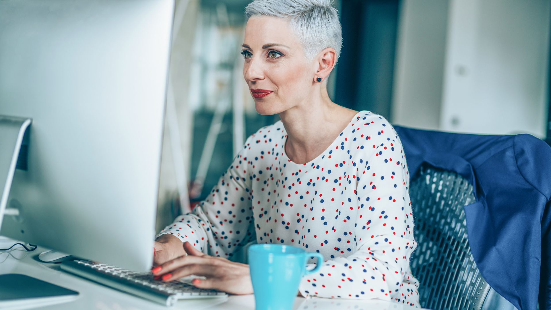 A woman with a short pixie cut and a polka dot top is using her computer in her office.
