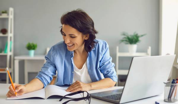 A woman is at her desk taking notes on her notebook while participating in a virtual training on her laptop.