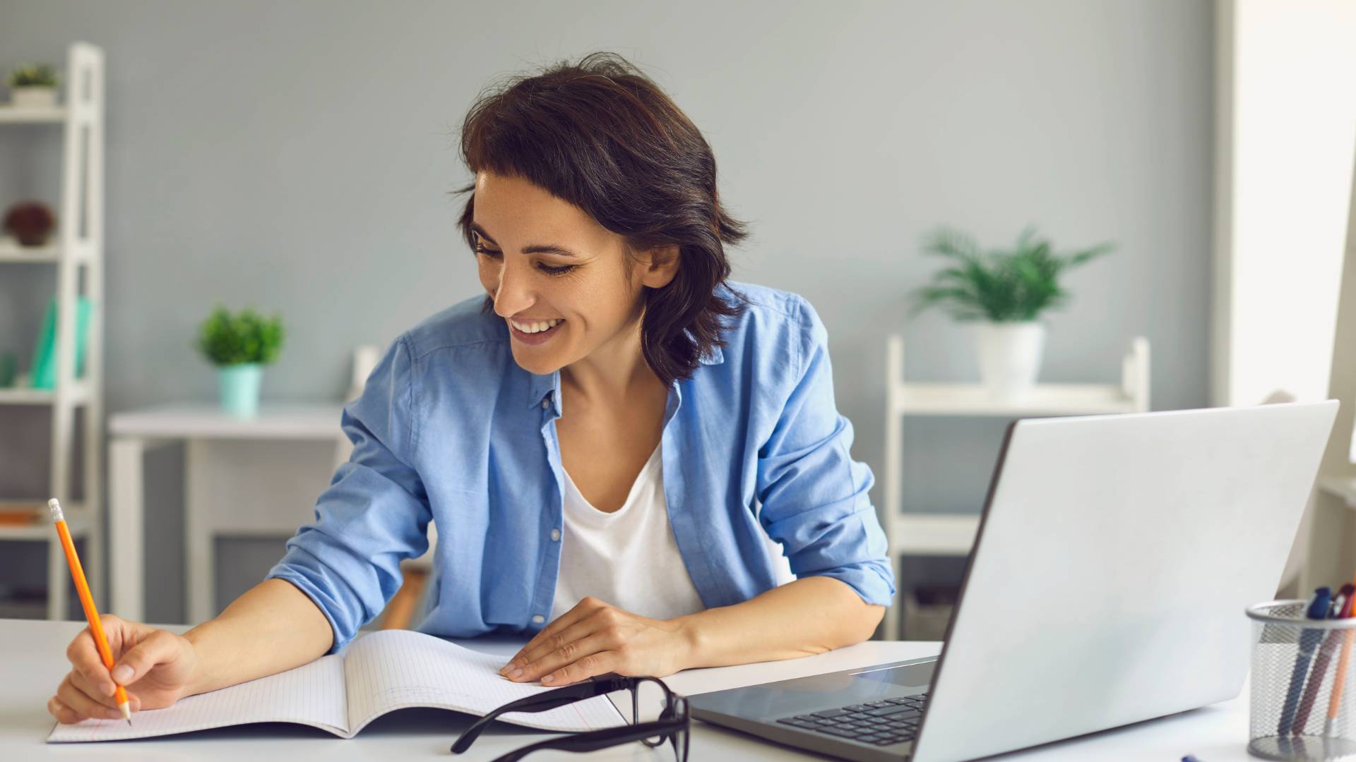 A woman is at her desk taking notes on her notebook while participating in a virtual training on her laptop.