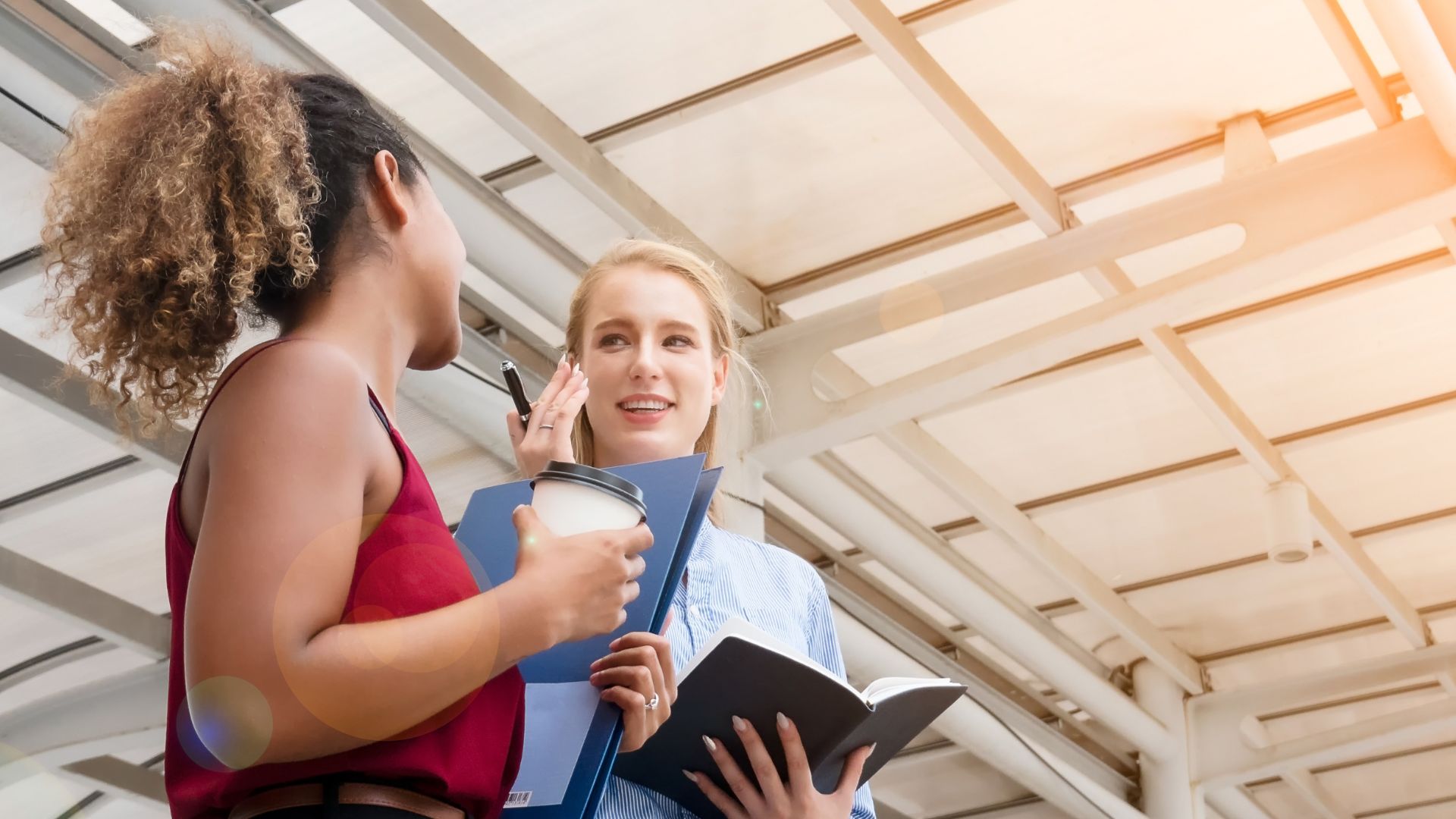 Two women engaging in a conversation outdoors under a shaded structure, exemplifying effective communication. One woman is holding a notebook and listening attentively while the other, holding a coffee cup, gestures as she speaks. They appear to be in a casual work-related discussion, highlighting an open, effective exchange of ideas.