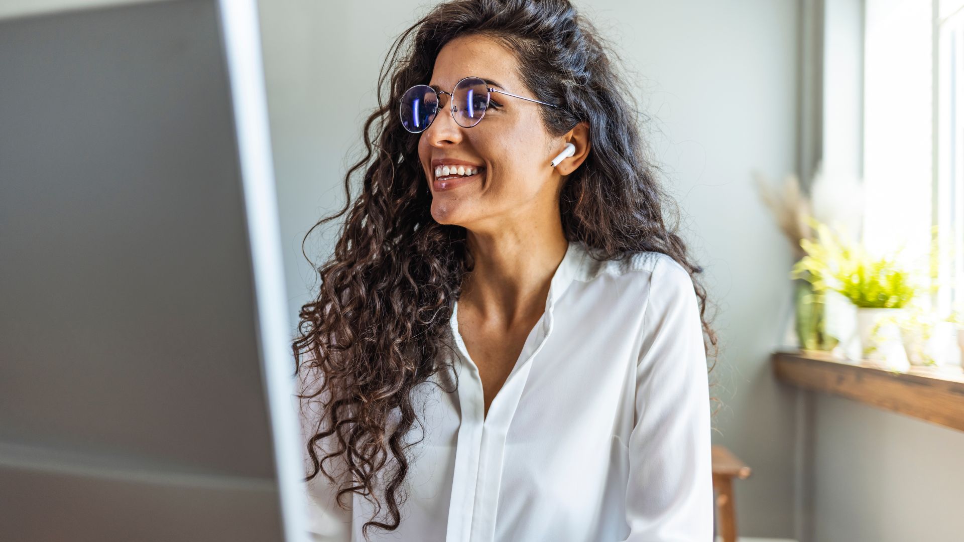 A woman is smiling at her monitor in her home office. She has her ear buds in.