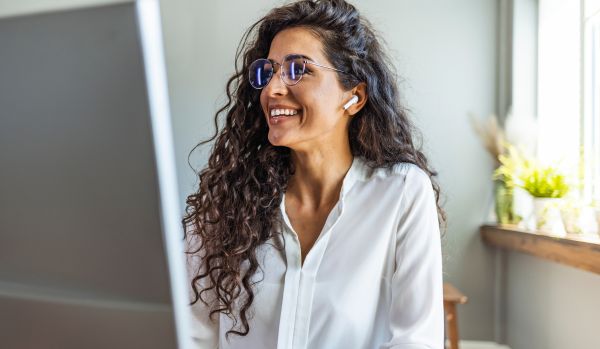 A woman is smiling at her monitor in her home office. She has her ear buds in.