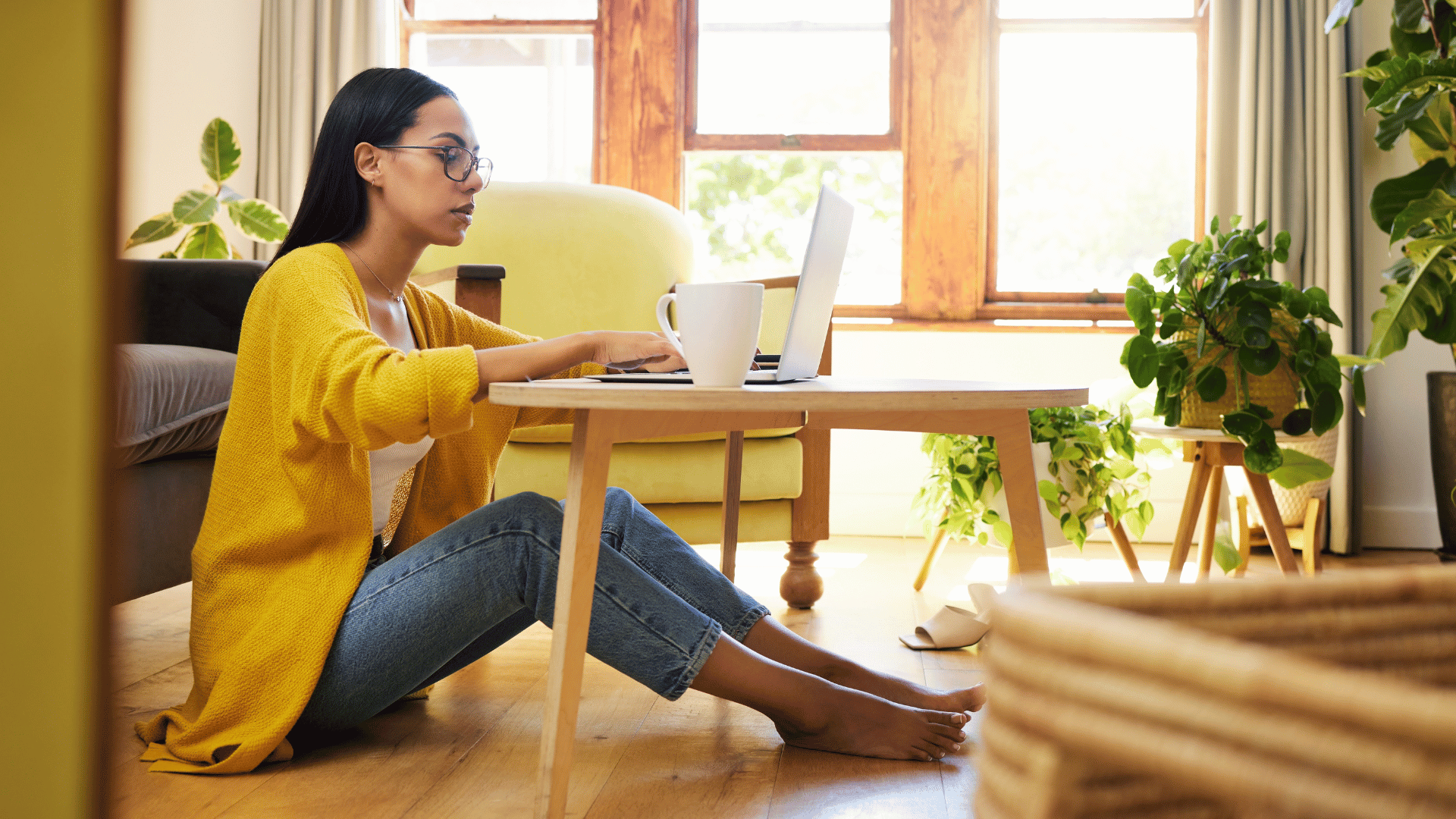 A woman working at her computer. She's sitting in front of a coffee table, with her legs under the coffee table. She has a cup of coffee next to her computer. She's wearing a yellow cardigan, white tanktop, jeans and glasses. She's barefoot and appears to be working from home.