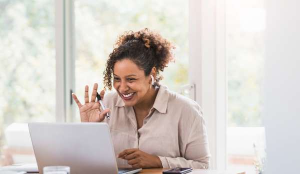 A middle-aged woman is smiling and waving at her laptop as she joins a live virtual training.