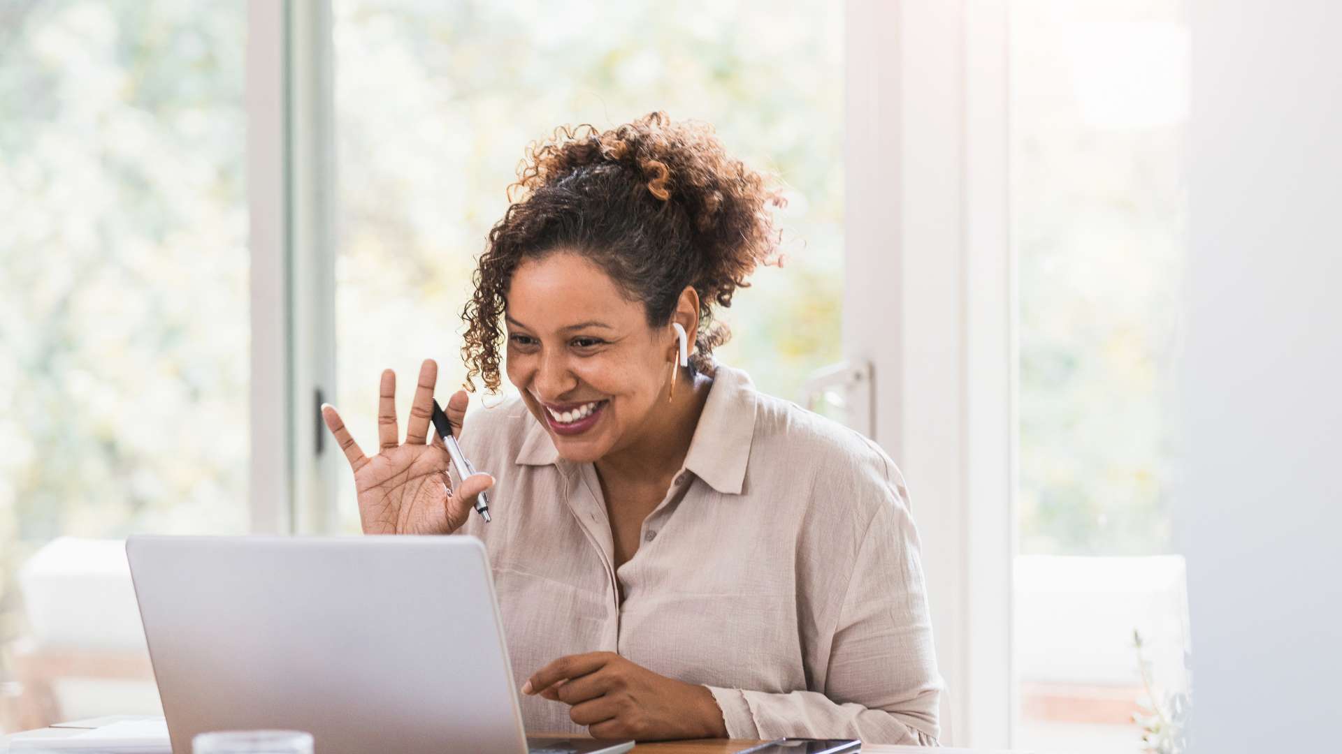 A middle-aged woman is smiling and waving at her laptop as she joins a live virtual training.