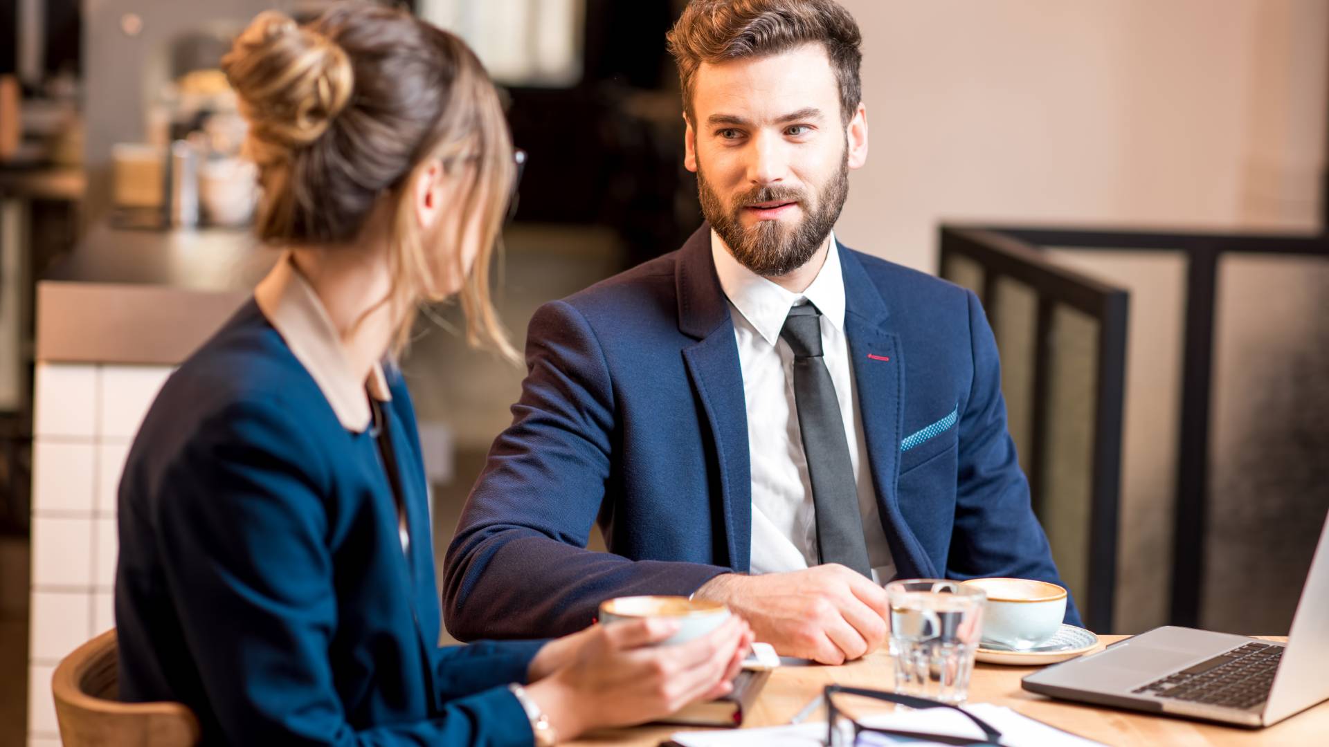 A man is having a feedback session with his female manager. We are looking over the woman's shoulder at the man. They are sitting at a table with coffee and their laptops.