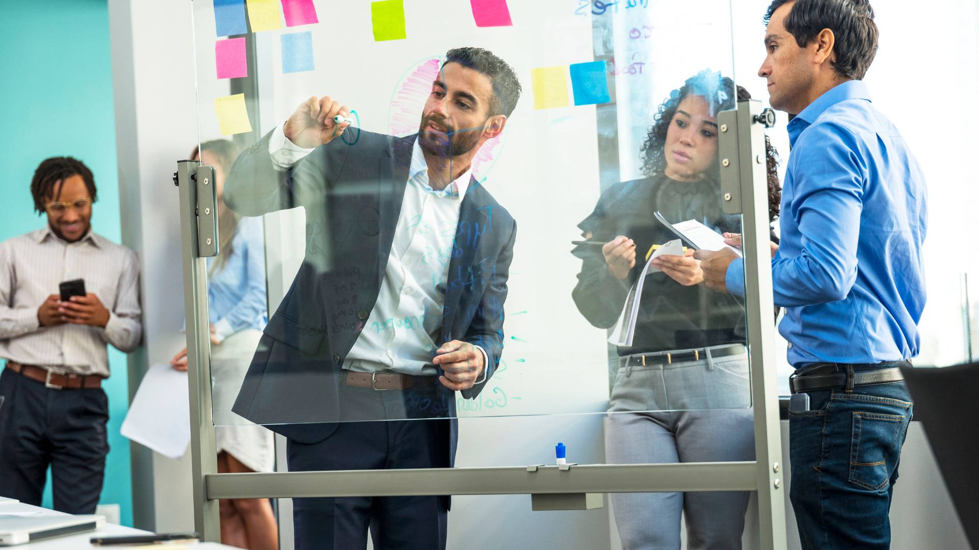 A man is drawing on a clear whiteboard while his team is watching and taking notes.