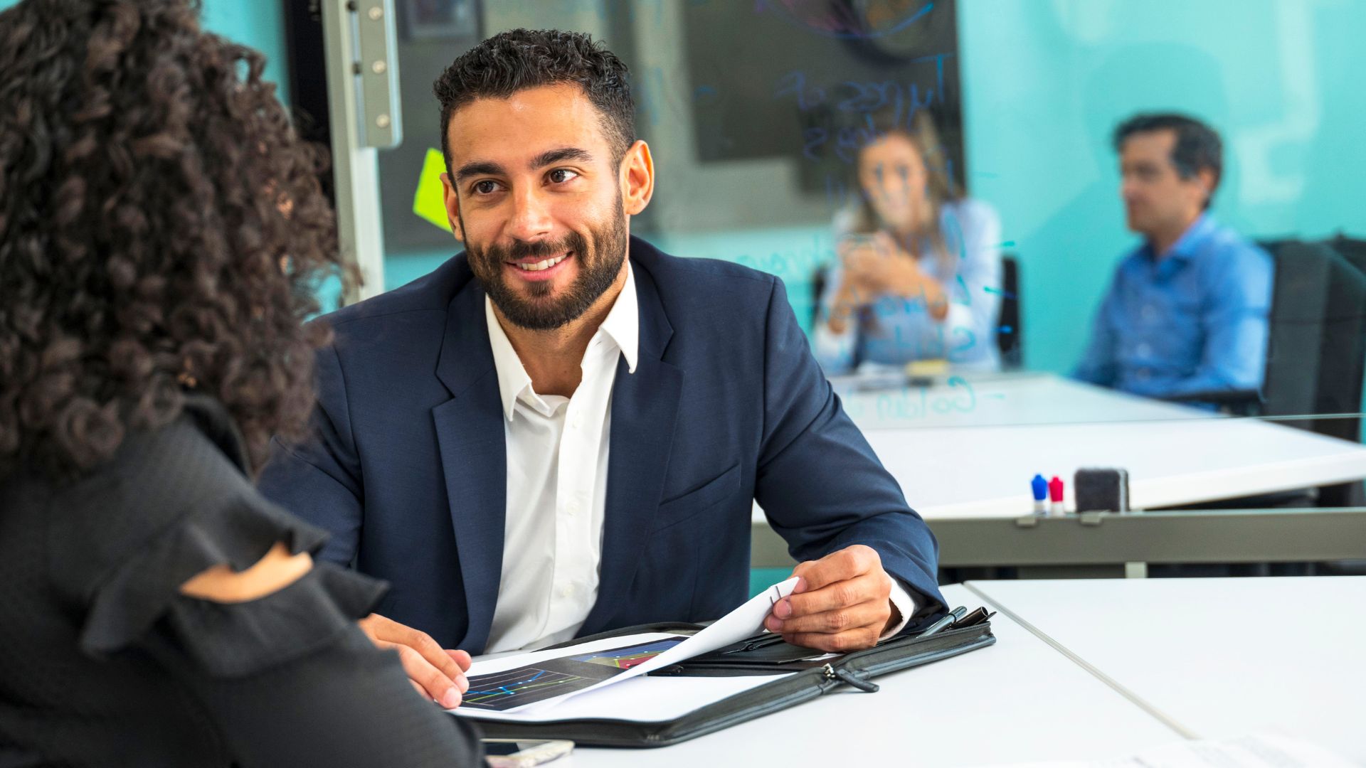 A man is meeting with a female-presenting colleague. We are looking over the woman's shoulder at the man. Two colleagues are visible in the background.