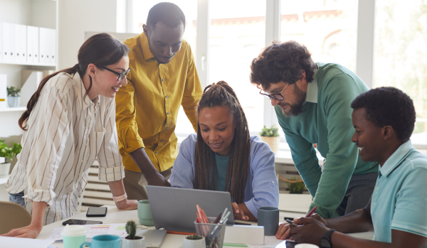 There are five people around a computer collaborating on a project. One woman is sitting down at the desk while everyone else is standing around her.