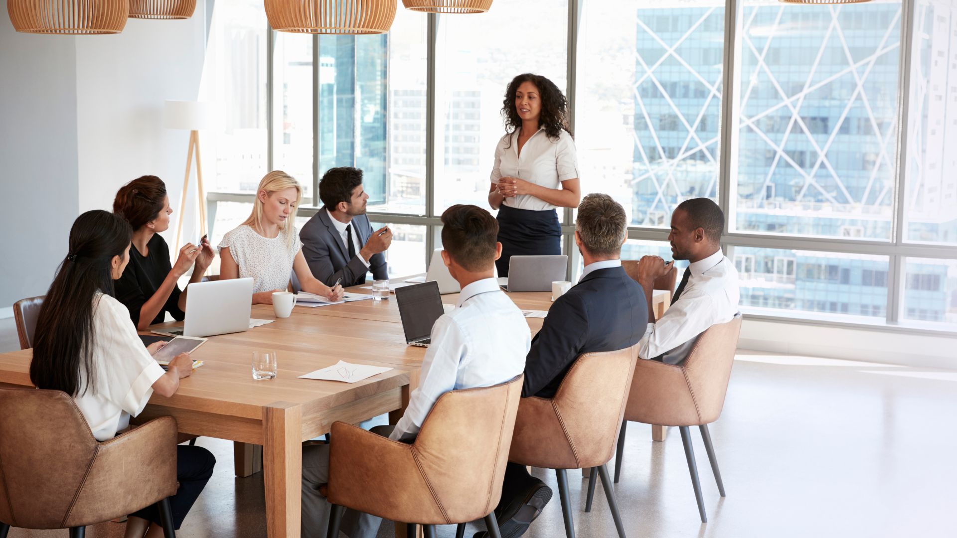 A woman is presenting to a group of diverse colleagues who are seated around a table.