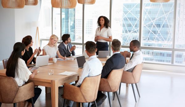 A woman is presenting to a group of diverse colleagues who are seated around a table.