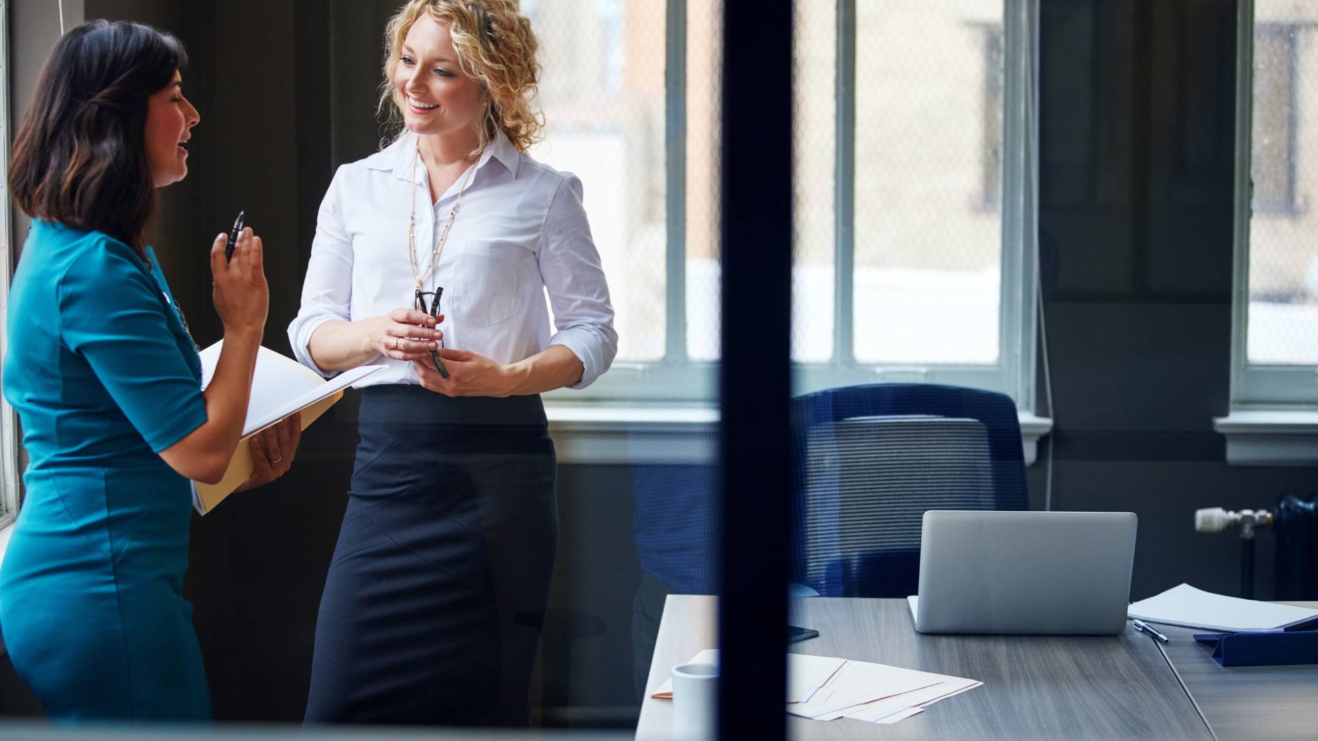 Two professional women are standing and having a conversation in an office.