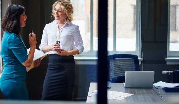 Two professional women are standing and having a conversation in an office.