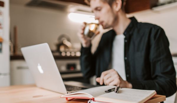 A man is taking a drink of water while working at his home desk with his laptop in front of him.