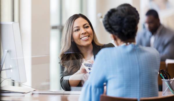 There's two women sitting down at a desk. One is Black and she's facing towards a Latina woman sitting on the other side of the desk.