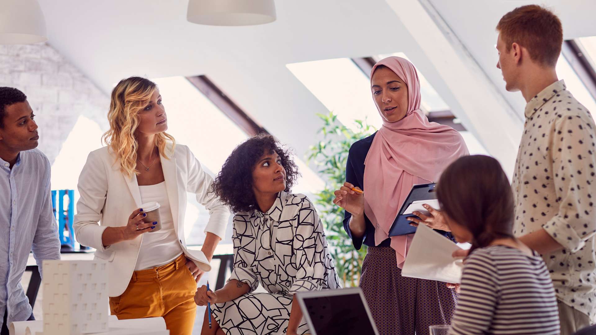 A diverse group of people are meeting in an office, listening to a woman in a pink hijab speak.