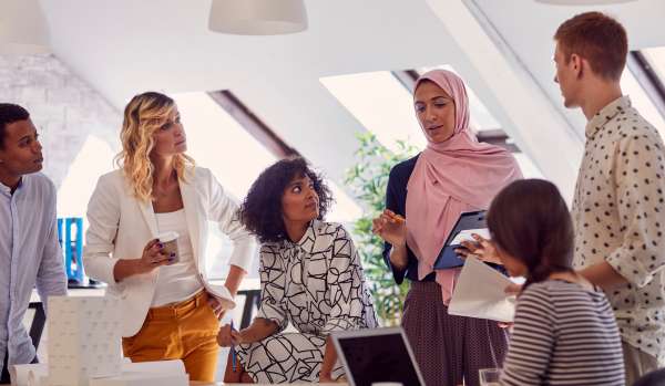 A diverse group of people are meeting in an office, listening to a woman in a pink hijab speak.