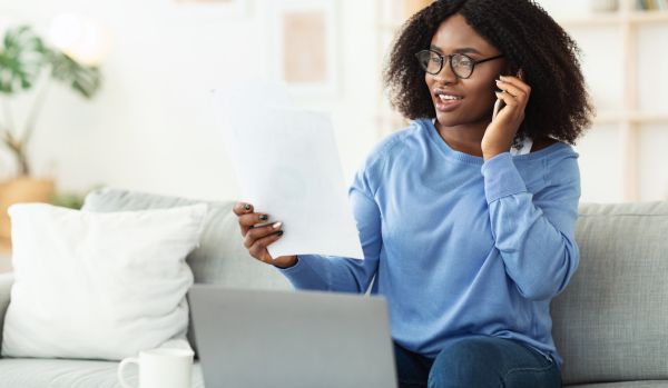 A young woman is working on her laptop on the couch while talking on the phone and holding a document.
