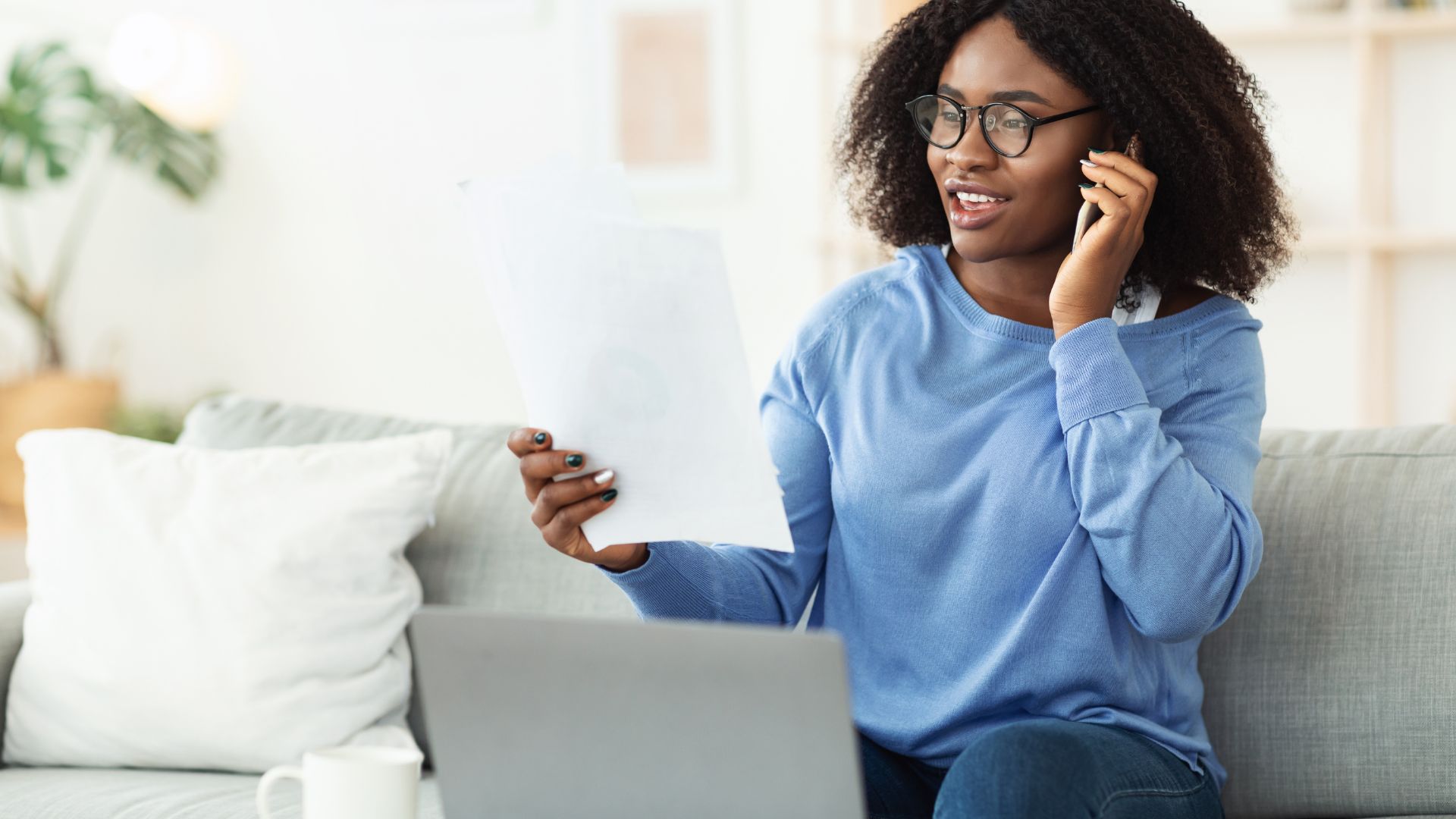 A young woman is working on her laptop on the couch while talking on the phone and holding a document.