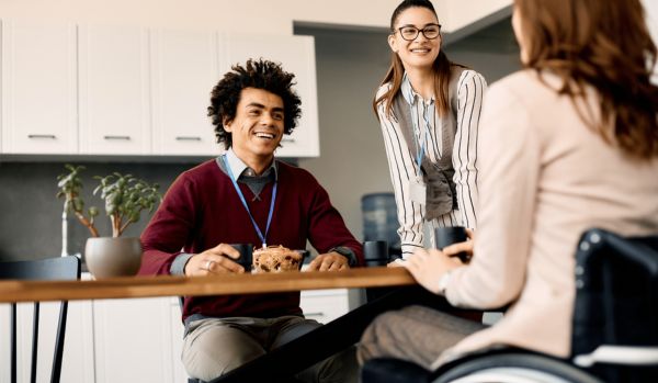 A diverse group of office workers are talking and smiling around a break room table.